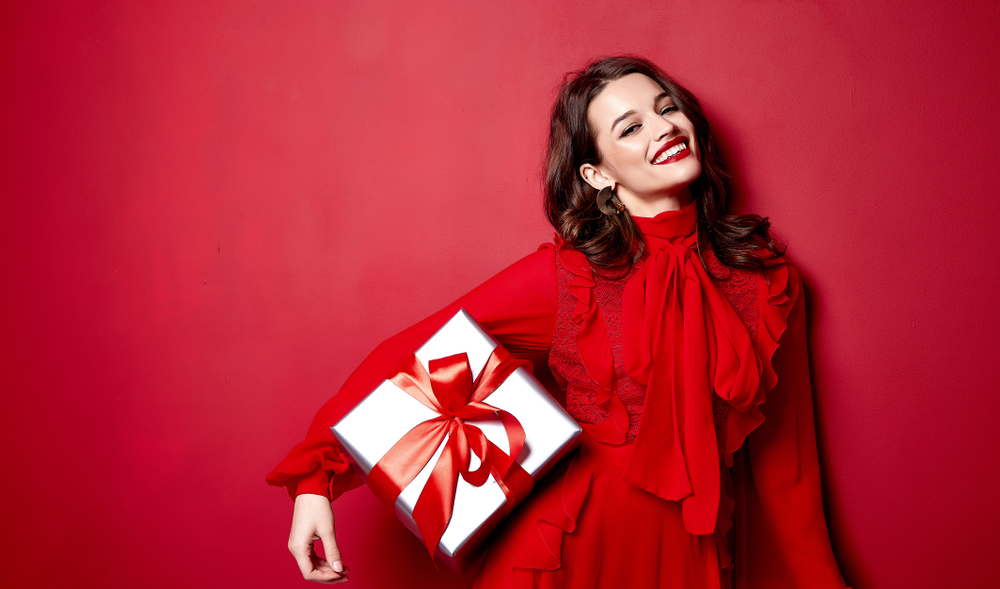 brown haired woman in a red frilled dress and red lipstick standing in front of a red background while holding a white present with a red ribbon in one arm and smiling